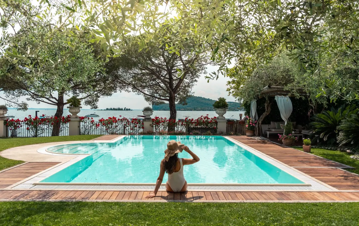 A guest in a hat sitting on the edge of the pool, among olive trees and pines, with the lake in the background